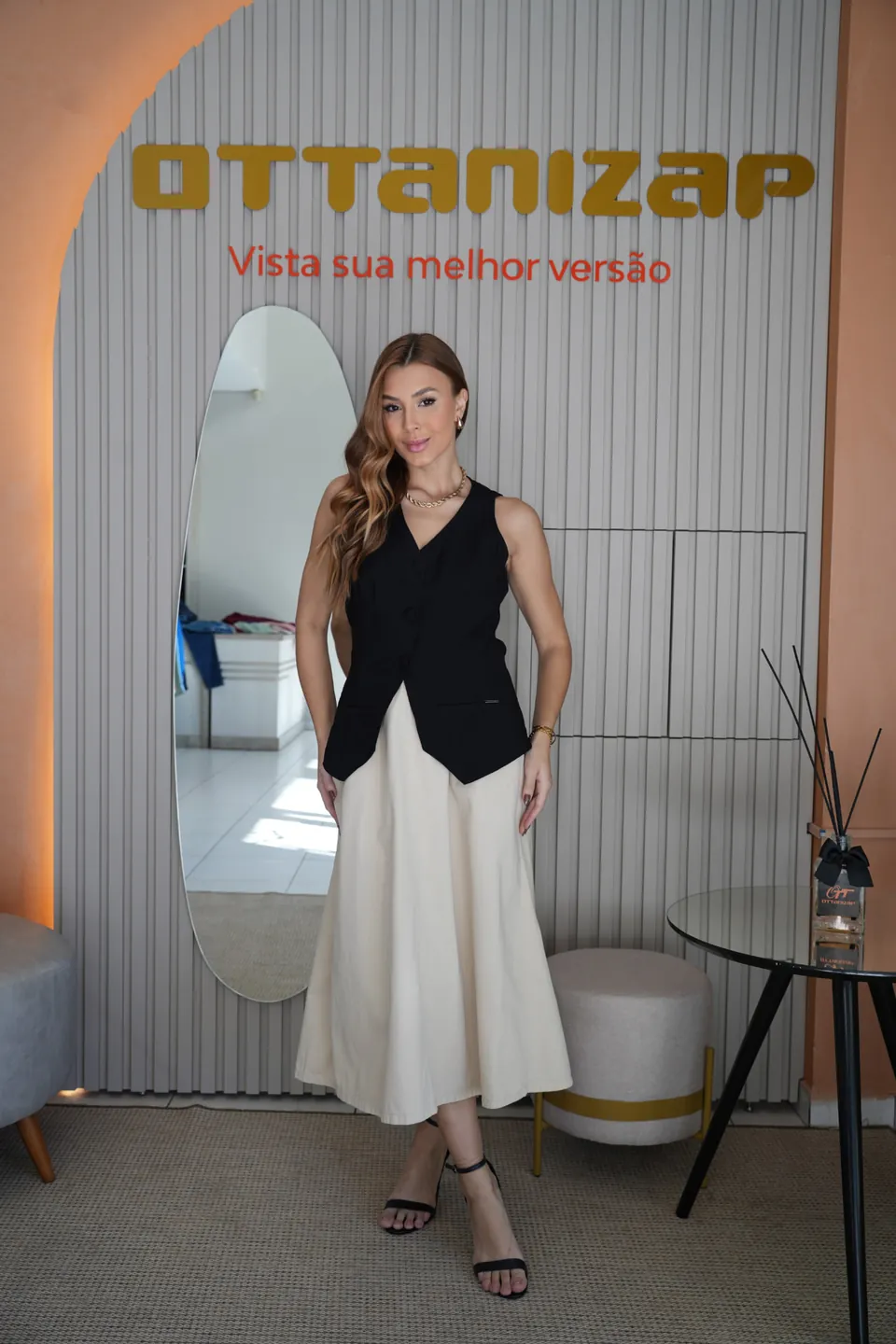 Modelo com colete preto e saia clara na Ottanizap Loja da Fábrica, diante da parede com a frase Vista sua melhor versão
